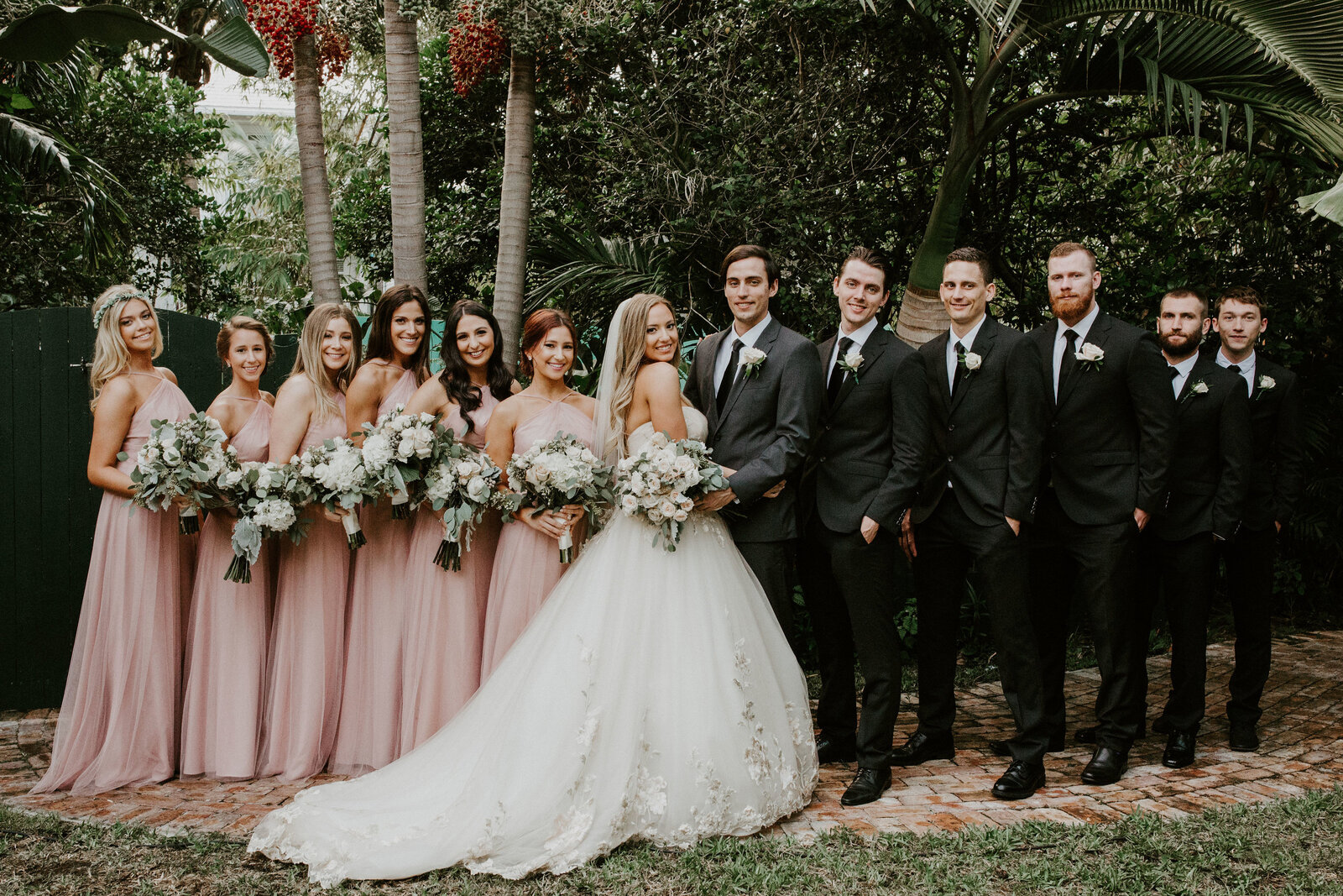 The full wedding party in black tie and blush gowns gathered beside the garden pond